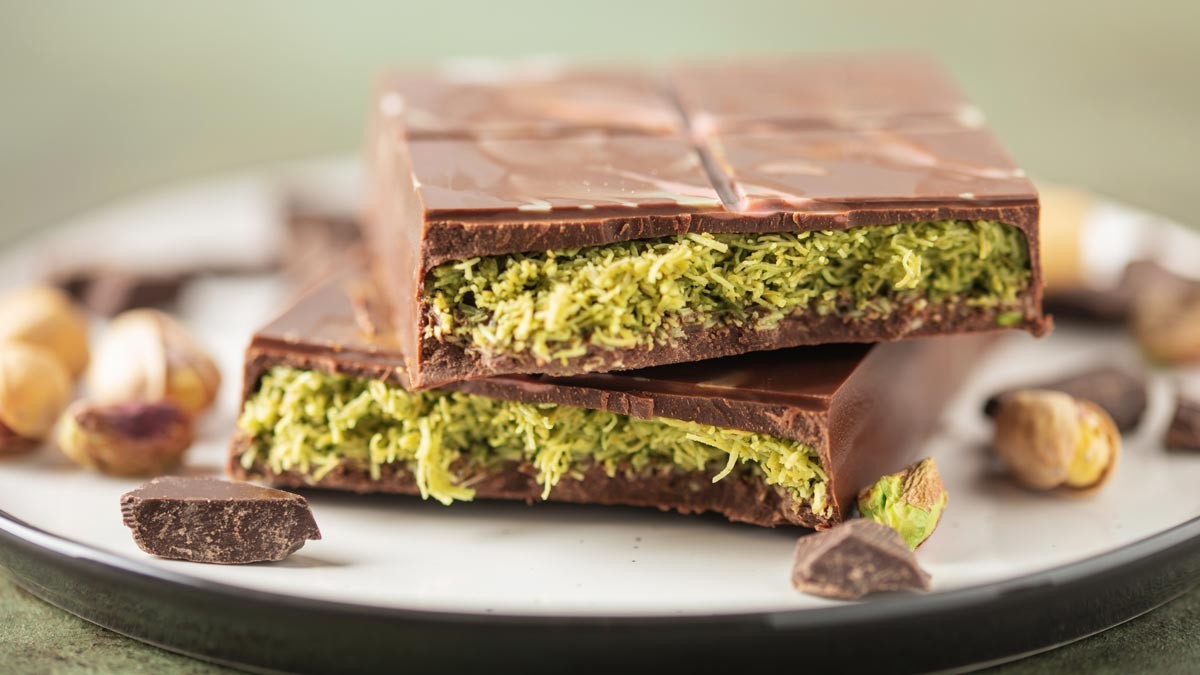 Two halves of a bar of Dubai chocolate lying on a white plate surrounded by pistachios and pieces of chocolate. Copyright: © murziknata / Adobe Stock
